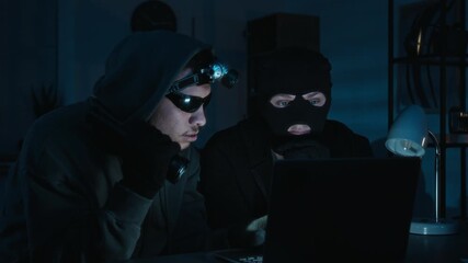 Two young male burglars looking at laptop screen studying confidential information when suddenly turning around upon hearing noise during late-night office break-in