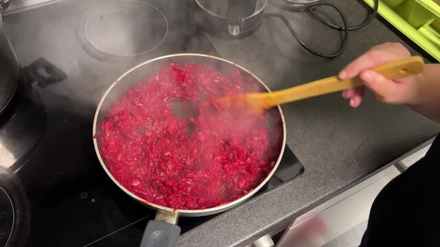 A close-up shot of shredded and sliced red beets and carrots being actively stewed and stirred in a frying pan on a black induction stove. The food is steaming and rich in color.