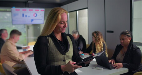 Businesswoman using digital tablet while team collaborates in background during office meeting, modern workplace with technology-driven productivity and professional focus