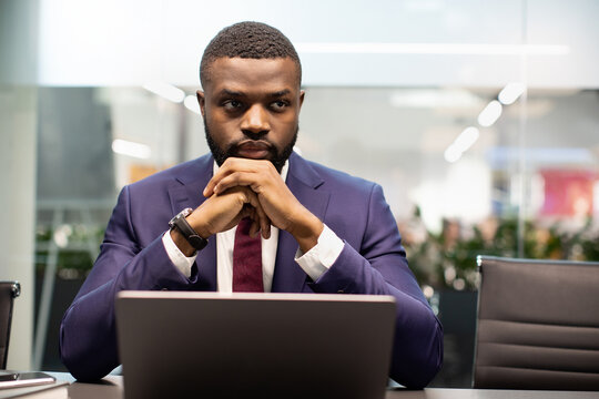 Frustrated young black businessman sitting at table in front of laptop at office, leaning on his hands, feeling depressed, needs for new business opportunities, looking at copy space