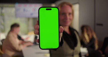 Close-up of businesswoman presenting smartphone with green screen in office meeting, diverse team working at conference table with analytics on screen
