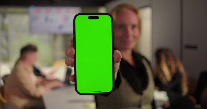 Close-up of businesswoman presenting smartphone with green screen in office meeting, diverse team working at conference table with analytics on screen