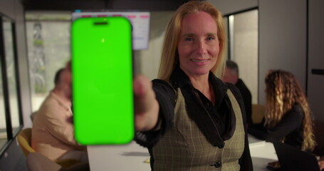 Close-up of businesswoman presenting smartphone with green screen in office meeting, diverse team working at conference table with analytics on screen