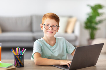 A young boy with red hair is sitting at a table, focused on his laptop while completing his homework. He enjoys online learning during the COVID-19 lockdown in a cozy home setting.