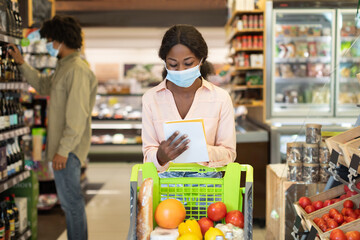 A woman in a pink shirt checks her shopping list while pushing a grocery cart filled with fresh...