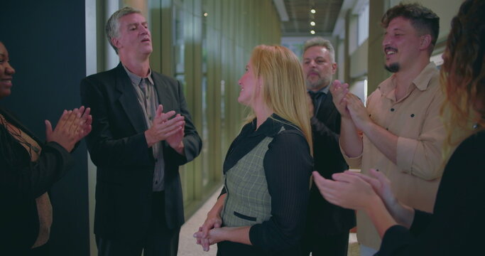 Cheerful business team clapping for female colleague in modern office hallway, diverse coworkers expressing recognition and appreciation for success