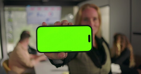 Close-up of hand holding smartphone with green screen, blurred business team working in modern conference room with charts on screen