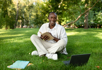 Obraz premium A man sits on the grass in a park, smiling while holding a book. A laptop and notebooks are nearby, creating a peaceful study setup under the trees.