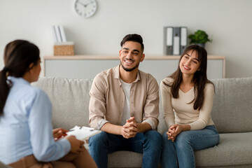A happy middle eastern couple sits on a couch at a counselor's office. They engage with the...