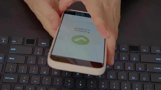 A woman holds a smartphone to complete a payment while sitting at a desk. The laptop keyboard is visible beneath her hands, highlighting modern transactions.