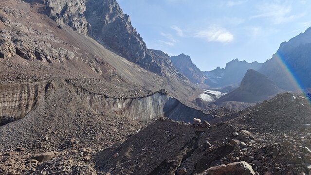 Glacial moraine and rugged mountain landscape with rainbow
High-altitude rocky terrain with visible glacier and layered moraine wall under clear sky and lens flare rainbow effect.