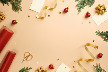 Christmas frame flat lay with red wrapping paper, gold ribbons, scissors, and baubles on beige...