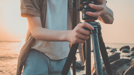 Person adjusting a camera tripod by rocky shoreline at sunset