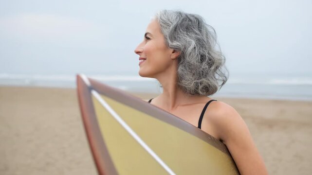 Mature woman gray hair swimsuit surfboard beach ocean smiling relaxed confident portrait joyful seaside lifestyle holding surfboard