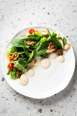 A vibrant chicken and spinach salad on a white plate, featuring walnuts, grapes, and a creamy dressing. The dish is presented close-up on a grey surface