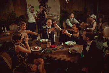 Stylish retro friends celebrating and toasting at festive dinner table setting. Concept of...