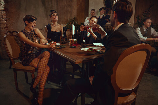 Vintage dinner party with stylish friends toasting and enjoying festive retro evening. Concept of holiday restaurant promos, gala dinner visuals, luxury event marketing, nightlife content sets.