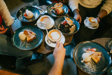 A group of people are sitting around a table with plates of food and drinks. There are several cups of coffee and tea, as well as a variety of desserts such as cakes and pies. The atmosphere is casual