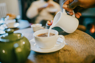 A person is pouring milk into a white cup of coffee. The scene is set in a cozy atmosphere with a wooden table and a few other cups and bowls on it