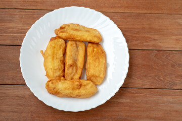 Overhead view of six golden fried bananas (Pisang Goreng) served in a paper-lined container on a rustic wooden table. Clean and simple Asian street food snack.