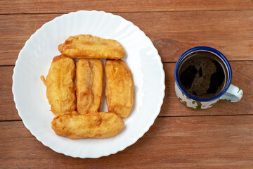 Overhead view of five fried bananas (Pisang Goreng) on a white fluted plate, served with a rustic enamel mug of black coffee on a wooden table.