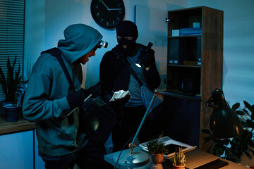 Two young adult men wearing masks and hoodies, stealing cash and valuables from office desk at...