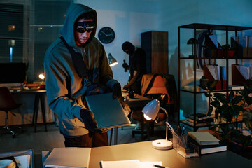 Young adult man wearing hood and mask stealing laptop from office desk while another masked man...