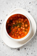 A close-up, appetizing photo of hearty red soup with sliced sausages, carrots, and herbs in a white bowl, perfect for food blogs or menus.