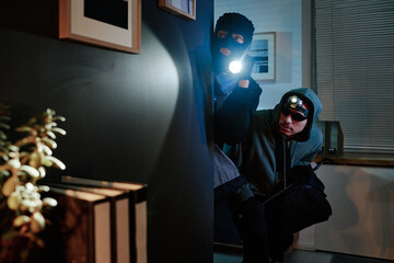 Two young adult men wearing masks and hoodies, sneaking through office holding flashlight and bag,...