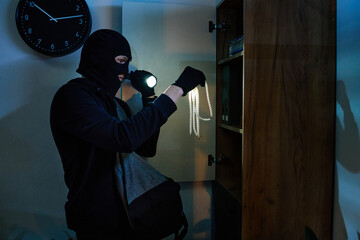 Young adult man wearing black balaclava and gloves holding flashlight and examining pearl necklace...