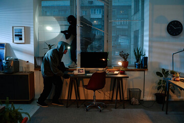 Two young adult men wearing masks and hoodies, breaking into office at night, one entering through...