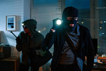 Two young adult men wearing masks and hoodies, breaking into office at night, holding flashlight...