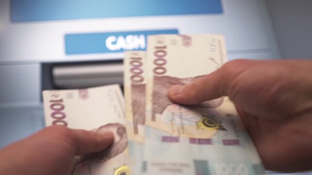 A close-up of a person's hand withdrawing money from an ATM. Ukrainian currency.