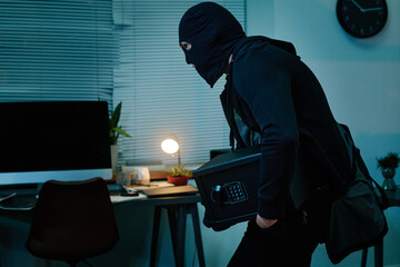 Middle aged man wearing black balaclava carrying electronic safe while sneaking through modern...