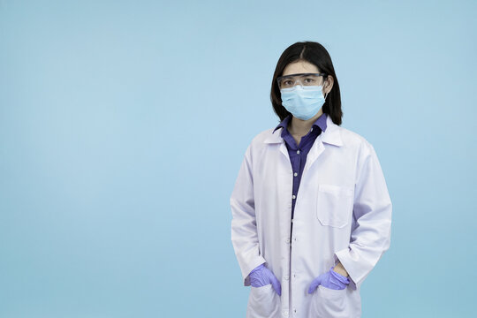 Confident Asian female scientist or doctor adjusting her lab coat and mask looking confidently in blue studio background