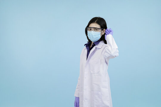 Confident Asian female scientist or doctor adjusting her lab coat and mask looking confidently in blue studio background