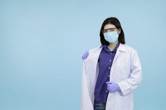 An Asian female scientist or doctor in a mask and safety glasses, adjusting her lab coat, symbolizing professional readiness and hygiene  in blue studio background