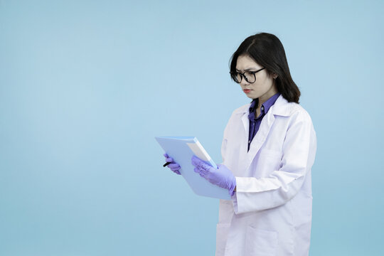 An Asian female scientist or doctor in a lab coat, diligently writing on a clipboard for data recording, research, or medical charting in blue studio background