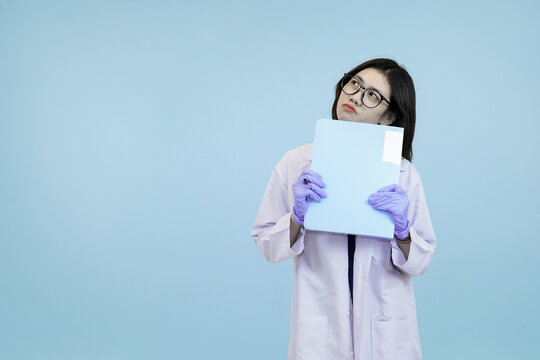 Female Asian scientist or doctor in a lab coat and gloves holding a blue folder that covers her face, symbolizing confidentiality or data privacy on a clear blue studio background - Powered by Adobe