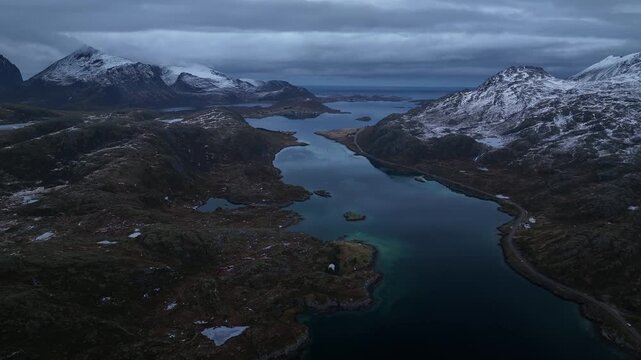 Aerial view of the dark blue waters winding between rugged, snow-dusted mountains under a moody sky, creating a dramatic contrast, Ballstad, Nordland, Norway.