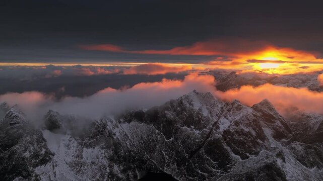 Aerial view of snow-covered mountain peaks kissed by the warm glow of the setting sun, creating a stunning contrast against the dark sky, Ballstad, Nordland, Norway.