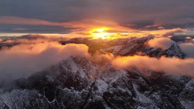 Aerial view of snow-capped mountain peaks piercing through a sea of clouds, bathed in the warm, ethereal glow of the setting sun, Ballstad, Nordland, Norway.