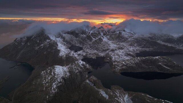 Aerial view of snow-capped mountains meeting the dark waters under a dramatic sky at sunset, Ballstad, Nordland, Norway.