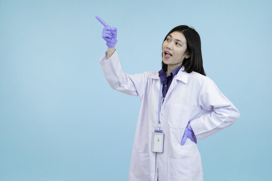 An assertive Asian female scientist or doctor in a lab coat and gloves, pointing to the side, signifying demonstration, presentation space in blue studio background