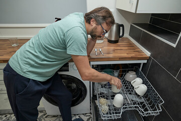 Man loading dishwasher in modern kitchen. Person doing household chores, cleaning dishes, and organizing kitchenware at home. Concept of domestic life, home cleaning, and modern lifestyle.
