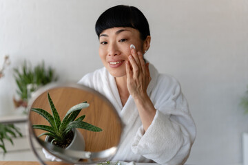 A smiling woman in a bathrobe applies skincare at a table with beauty products and plants, enjoying a calm self-care routine in natural light