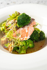 Close-up of a gourmet plated dish featuring pan-seared salmon fillet, broccoli florets, romanesco, fresh sprouts, and clams in a savory broth served in a white bowl.
