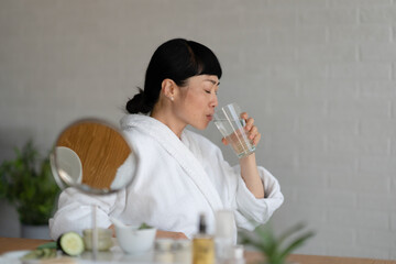 Asian woman in a white bathrobe drinking a glass of water at a table with skincare products, creating a calm and healthy self-care moment