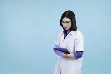 Focused Asian female scientist or doctor wearlab coat, writing data on a digital tablet, symbolizing meticulous research and modern record-keeping in blue studio background