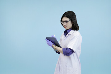 Focused Asian female scientist or doctor wearlab coat, writing data on a digital tablet, symbolizing meticulous research and modern record-keeping in blue studio background